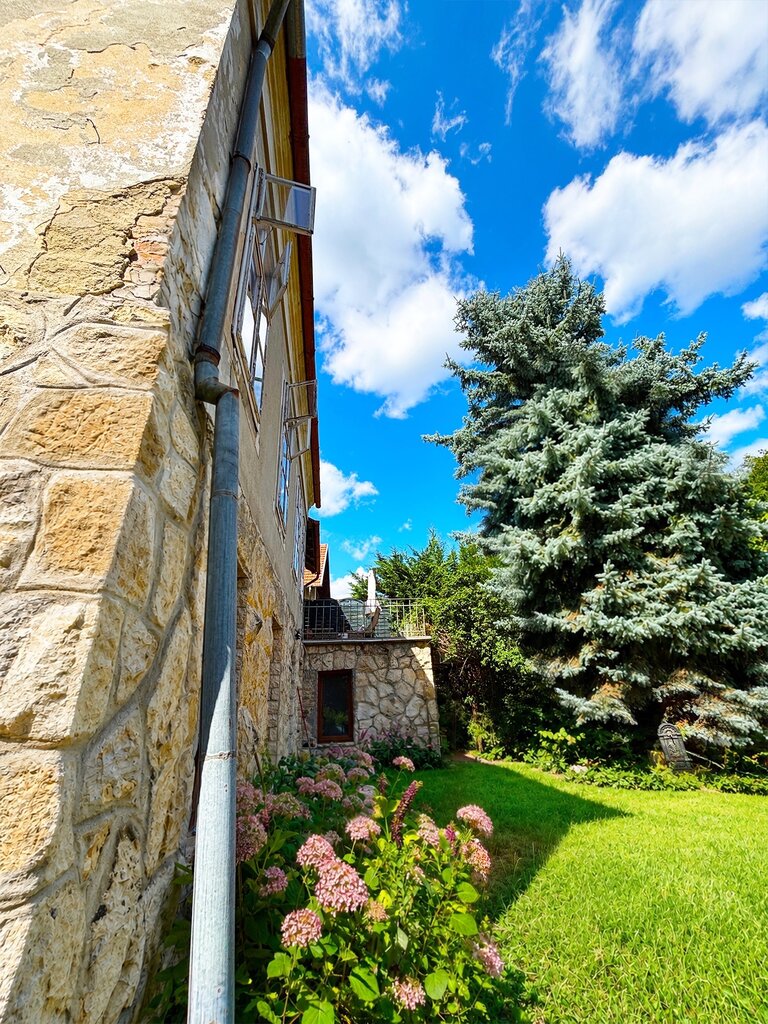 A villa homlokzata kő alapzattal, erkéllyel és nagy fenyőfával a kertben