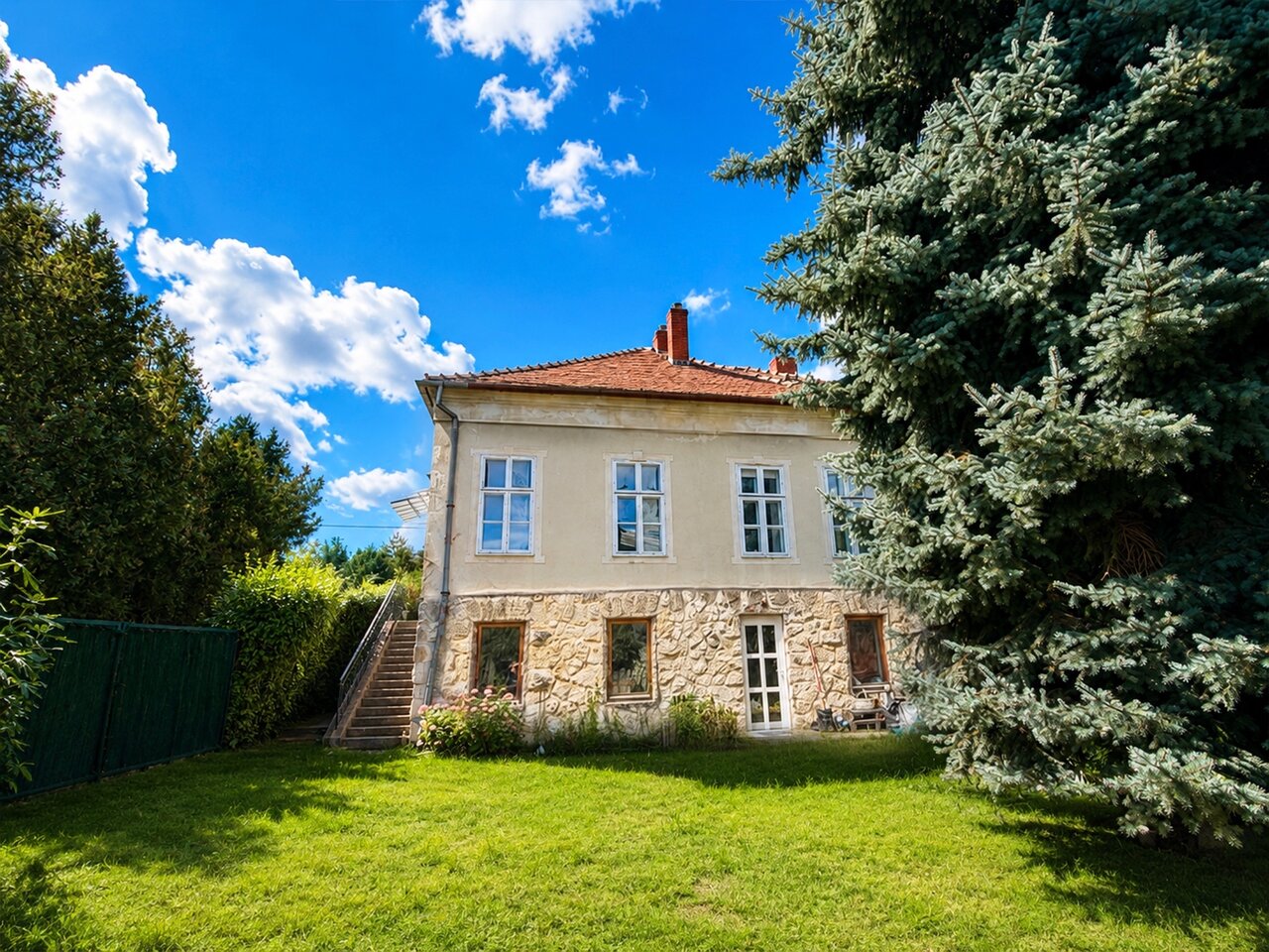 A budakeszi szecessziós villa homlokzata kőburkolatos alsó szinttel, gondozott kerttel és nagy fenyőfákkal